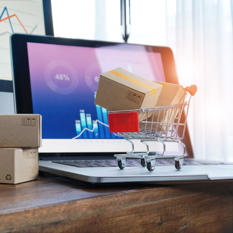 Laptop, open with finance stats on the screen, surrounded by small boxes (ecommerce packages) and a small trolley (to represent a shopping cart)